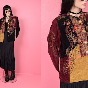 Op de afbeelding: Een vintage jasje met een patchwork ontwerp. De jas heeft paisley en bloemenborduurwerk in bordeauxrood, zwart en goud. Het heeft gouden knopen en een zwarte kraag. De jas wordt gedragen over een zwarte jurk en rok.