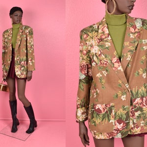 May include: A tan blazer with a floral pattern featuring pink, red, and white roses and green leaves. The blazer has two front pockets and a two-button closure. The model is wearing a green turtleneck and a burgundy skirt.