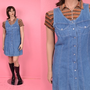 Puede incluir: Vestido vaquero azul claro con botones en la parte delantera y cuello en V. El vestido está superpuesto a una camisa de manga corta con cuello y un estampado de rayas marrones, naranjas y blancas. El vestido llega a la rodilla.