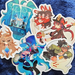May include: A collection of colorful anime-style stickers featuring various characters. The stickers showcase vibrant designs with characters in dynamic poses, including one with the text "Miku". The stickers are on a blue fabric surface.
