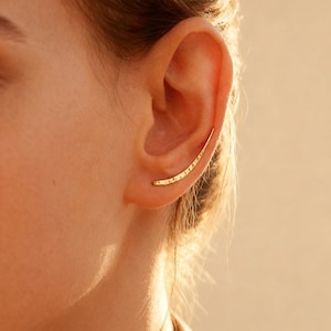 Peut inclure: Un ear cuff doré avec une texture martelée, incurvé pour suivre la forme de l'oreille. Le bijou est porté sur l'oreille, avec un fond chaud et neutre.