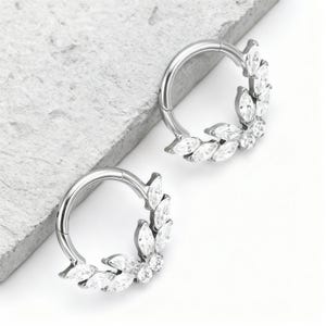 Pode incluir: Dois brincos de argola prateados. Cada brinco apresenta um design decorativo de cristais transparentes com corte marquesa e redondos, dispostos em forma de folha. O fundo é cinza claro e branco.