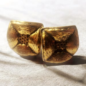 May include: A pair of gold-toned stud earrings with a square, slightly curved shape. The surface of each earring features a textured, hammered finish with a recessed center point that radiates outward toward the four rounded corners.