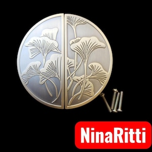 Puede incluir: Un tirador de puerta circular de metal dividido en dos mitades, con acabado en plata cepillada y un patrón de hojas de ginkgo en relieve. El juego incluye tres pequeños tornillos de montaje. El texto NinaRitti aparece en un recuadro rojo.