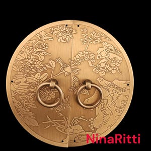 May include: A circular brass door plate featuring an engraved nature scene with birds, butterflies, and floral motifs. The plate is split into two halves, each equipped with a circular ring handle. The text NinaRitti is printed in red.