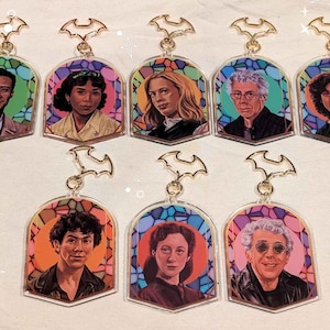 Pode incluir: Chaveiros de acrílico com retratos de pessoas em um design de vitral. Cada chaveiro tem um topo dourado em forma de morcego. Os retratos são colocados em um fundo colorido de laranja, azul e roxo.