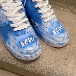 May include: A pair of low-top sneakers featuring a light blue and white cloud pattern across the leather upper. The toe box of each shoe displays the white text "KEVA" in a stylized font, complemented by white laces and a white rubber sole.