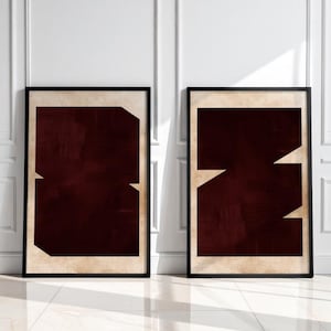May include: Two framed abstract art prints. Each print features a dark burgundy geometric design with beige accents, set against a beige background. The frames are black, and the artwork is displayed against a white wall and a glossy white floor.