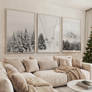 May include: Three framed monochrome prints showing snowy mountain scenes and forests. They hang above a beige sectional sofa with cushions and a knitted throw. A Christmas tree is visible on the right.