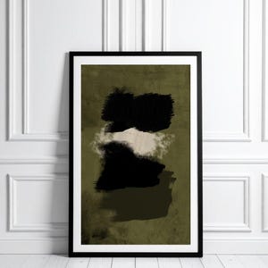 May include: Abstract art print in a black frame. The artwork features black brushstrokes over a muted olive green background, with a white and beige cloud-like shape in the middle. The framed art is displayed against a white wall and wooden floor.