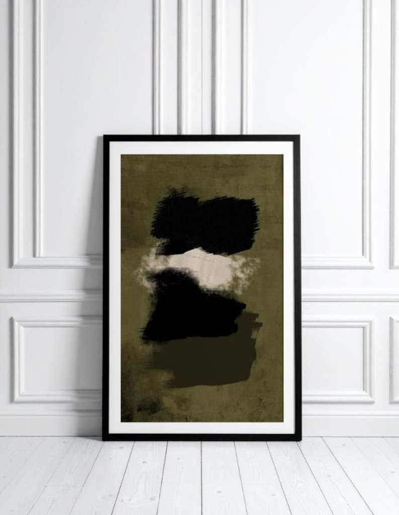 May include: Abstract art print in a black frame. The artwork features black brushstrokes over a muted olive green background, with a white and beige cloud-like shape in the middle. The framed art is displayed against a white wall and wooden floor.
