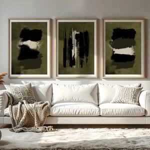 May include: Three framed abstract art prints. Each print features a dark olive green background with black and white brushstroke designs. The prints are displayed above a white sofa with decorative pillows and a neutral-toned throw blanket.
