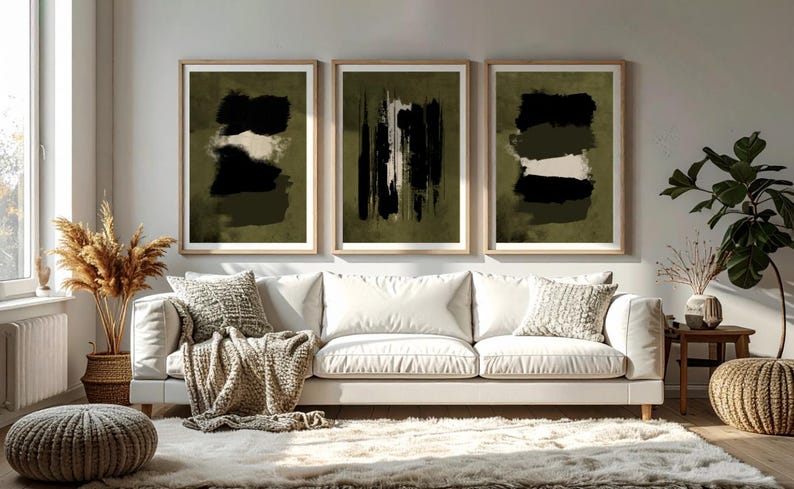 May include: Three framed abstract art prints. Each print features a dark olive green background with black and white brushstroke designs. The prints are displayed above a white sofa with decorative pillows and a neutral-toned throw blanket.