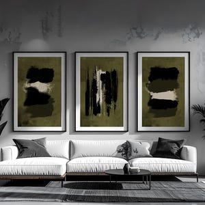 May include: Three framed abstract art prints. Each print features a black and white design on an olive green background. The prints are displayed above a white sofa with black pillows.