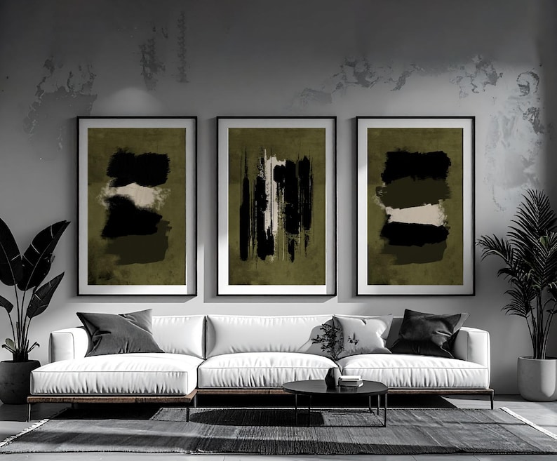 May include: Three framed abstract art prints. Each print features a black and white design on an olive green background. The prints are displayed above a white sofa with black pillows.