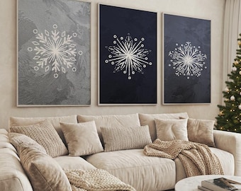 Könnte beinhalten: Drei gerahmte Wandkunstwerke mit weißen Schneeflockenmustern auf strukturierten Hintergründen in Grau, Marineblau und Dunkelgrau. Die Kunstwerke hängen über einem beigefarbenen Ecksofa mit dekorativen Kissen und einer Strickdecke.