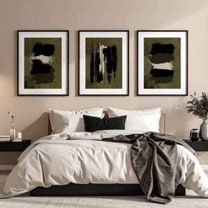 May include: Three framed abstract art prints with black frames hang above a bed. The art features brushstroke designs in black, white, and olive green. The bed has white bedding, black pillows, and a gray throw blanket. Black nightstands flank the bed.
