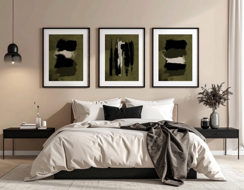 May include: Three framed abstract art prints with black frames hang above a bed. The art features brushstroke designs in black, white, and olive green. The bed has white bedding, black pillows, and a gray throw blanket. Black nightstands flank the bed.