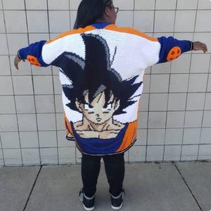 Può includere: Cardigan all'uncinetto fatto a mano con una grande grafica di un personaggio della serie anime Dragon Ball Z. Il cardigan ha un corpo bianco con dettagli arancioni, blu e neri. Il volto del personaggio è ben visibile sul retro.