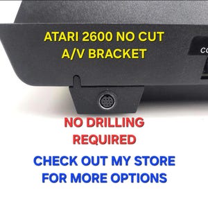 May include: A black Atari 2600 no cut A/V bracket featuring a circular port. The text on the image reads: ATARI 2600 NO CUT A/V BRACKET, NO DRILLING REQUIRED, CHECK OUT MY STORE FOR MORE OPTIONS. The bracket is designed for a game console.