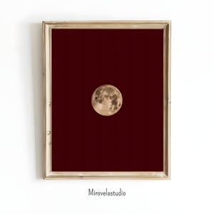 Puede incluir: Impresión enmarcada que presenta una luna llena sobre un fondo rojo oscuro. La luna está detallada con cráteres y diferentes tonos de gris y marrón. El marco es de color dorado metálico, y la impresión está sobre un fondo blanco. El texto "Mirovelastudio" está en la parte inferior.