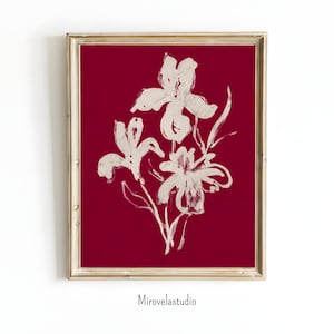 Puede incluir: Una impresión artística enmarcada con una ilustración al estilo acuarela de flores de iris blancas sobre un fondo rojo intenso. La obra de arte está en un marco dorado e incluye el texto "Mirovelastudio" en la parte inferior.