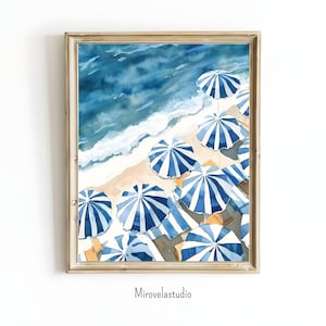 Puede incluir: Pintura a la acuarela de una escena de playa con sombrillas a rayas azules y blancas, orilla arenosa y olas del océano. La obra de arte está enmarcada en un marco dorado. El nombre del artista, Mirovelastudio, está impreso en la parte inferior.