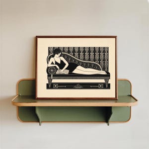 Peut inclure: Une estampe Art déco en noir et blanc encadrée, représentant une personne lisant sur une chaise longue. L'estampe est exposée sur une étagère verte et boisée, fixée à un mur gris clair. L'œuvre présente des motifs géométriques.