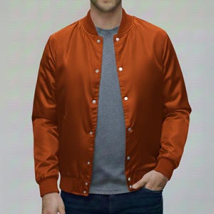 Puede incluir: Chaqueta bomber naranja quemado con cuello y puños de canalé. La chaqueta tiene cierre de botones a presión y un acabado satinado. El modelo lleva una camiseta gris y vaqueros azules. La chaqueta es una prenda de abrigo informal.