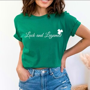 May include: A white ceramic mug with a black handle and the text "But First, Coffee" in a bold, black font. The mug is set against a blurred background. The mug appears to be approximately 10 cm tall.