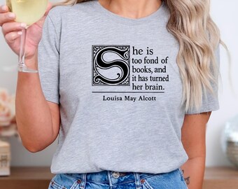 Può includere: T-shirt grigio chiaro con la citazione "he is too fond of books, and it has turned her brain." di Louisa May Alcott. La citazione è in nero, con una "S" decorativa. La maglietta è abbinata a jeans blu.