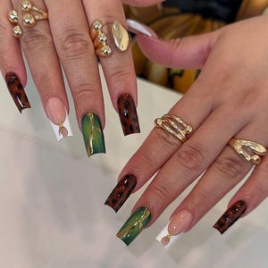 May include: Close-up of hands with long, square-shaped acrylic nails. The nails feature various designs, including a tortoiseshell pattern, green and gold accents, and a French tip with a gold charm. Gold rings adorn the fingers.