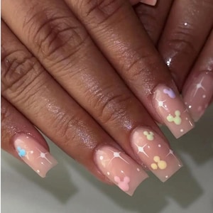 May include: Square-shaped acrylic nails with a light pink base. The nails feature white starburst designs and pastel-colored Mickey Mouse head designs. The nails are on a hand with dark skin.