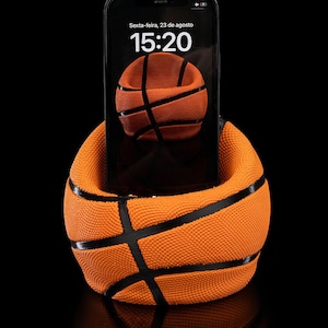 May include: An orange basketball-themed phone holder with black lines, holding a black smartphone. The phone displays a basketball image on the screen, with the time and date visible. The holder sits on a reflective black surface.