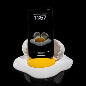 Può includere: Uno smartphone nero con un'immagine di uovo rotto, appoggiato su un supporto in ceramica bianca a forma di uovo con un tuorlo giallo. Lo schermo del telefono mostra l'ora 11:57 e la data 1 agosto. Lo sfondo è nero.