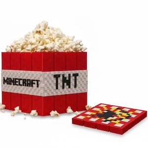 Peut inclure: Un récipient à popcorn rouge sur le thème de TNT rempli de pop-corn, avec les mots "MINECRAFT" et "TNT" sur le côté. Un bloc TNT rouge et noir assorti est à côté du récipient. Des grains de pop-corn sont éparpillés.
