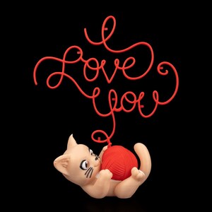 Puede incluir: Figura de gato beige jugando con una bola de hilo roja, con las palabras "I Love You" en rojo arriba. El fondo es negro.