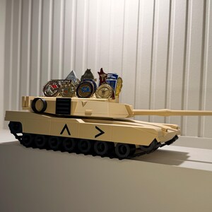 May include: Tan toy tank with black treads and a long barrel. Several collectible coins and small figurines are displayed on top of the tank. The tank has black chevrons on the side.