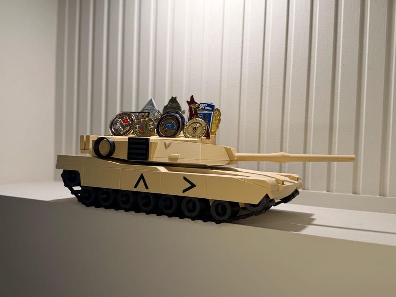 May include: Tan toy tank with black treads and a long barrel. Several collectible coins and small figurines are displayed on top of the tank. The tank has black chevrons on the side.