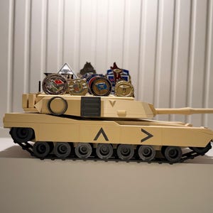 May include: A tan toy tank with black treads and a long barrel. Several military-style coins and emblems are displayed on top of the tank. The tank has black chevron markings on the side.
