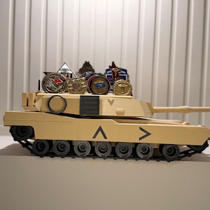 May include: A tan toy tank with black treads and wheels. Several military-style pins and medallions are displayed on top of the tank. The tank has a long barrel and directional arrows.