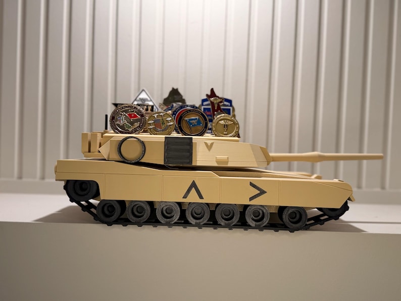May include: A tan toy tank with black treads and wheels. Several military-style pins and medallions are displayed on top of the tank. The tank has a long barrel and directional arrows.