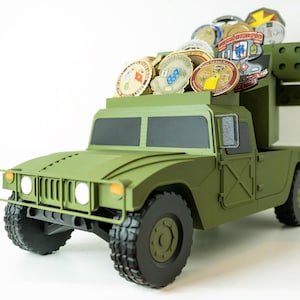 May include: An olive green military-style truck model featuring a flatbed cargo area filled with various circular metal challenge coins. The vehicle has black tires, a front grille guard, and a boxy frame design with dark windows and doors.