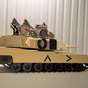 May include: A tan-colored toy tank with black treads and wheels. The tank has a series of coins and a small plaque on top. The tank has a black circle and a black rectangle on the top. The tank has a chevron symbol on the side.