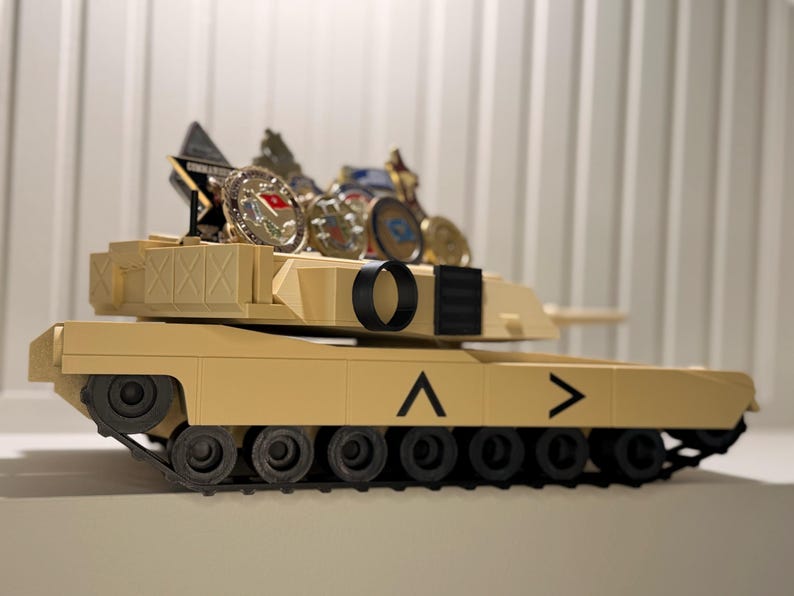 May include: A tan-colored toy tank with black treads and wheels. The tank has a series of coins and a small plaque on top. The tank has a black circle and a black rectangle on the top. The tank has a chevron symbol on the side.