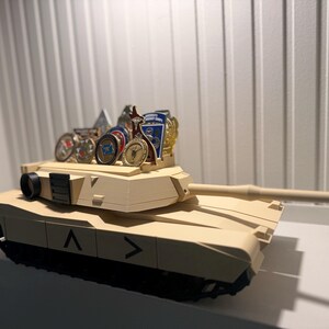 May include: A tan-colored toy tank with a long barrel and black treads. The tank has a series of military-style medals and pins displayed on top. The tank is sitting on a white surface.
