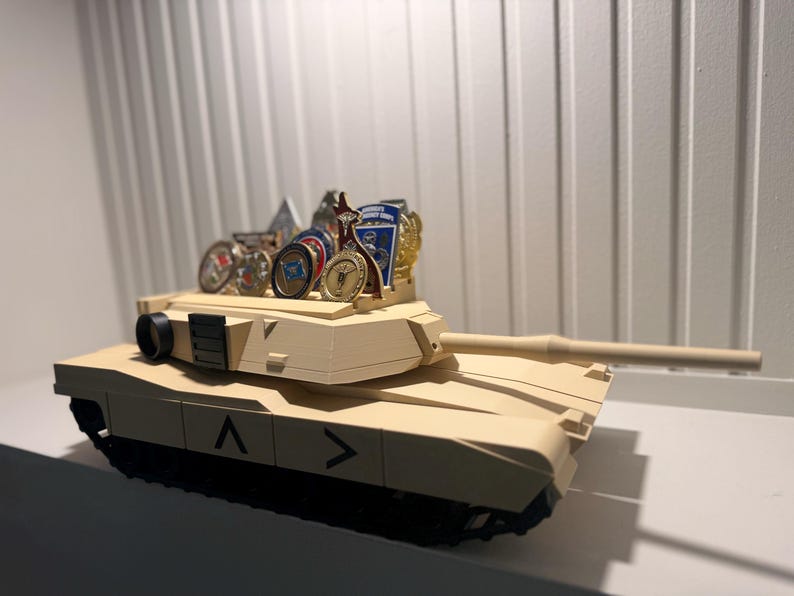 May include: A tan-colored toy tank with a long barrel and black treads. The tank has a series of military-style medals and pins displayed on top. The tank is sitting on a white surface.