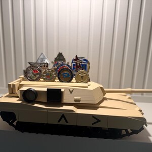 May include: A tan toy tank with black treads and a long barrel. Several military challenge coins are displayed on top of the tank. The tank has black markings and a black circular object on the side.