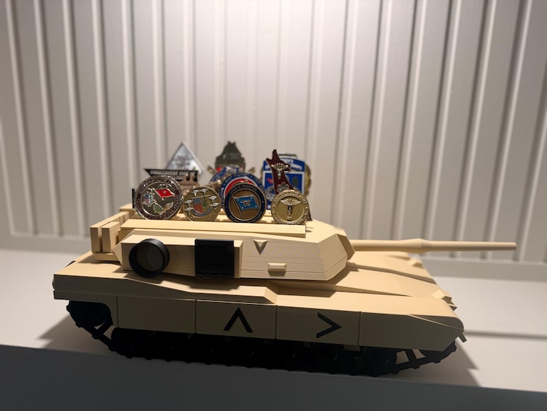 May include: A tan toy tank with black treads and a long barrel. Several military challenge coins are displayed on top of the tank. The tank has black markings and a black circular object on the side.