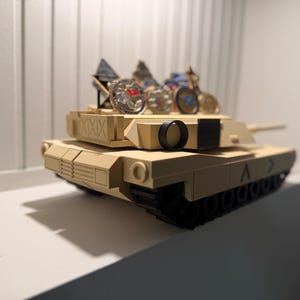May include: A tan toy tank with black treads and a black circular detail. Several small, decorative items are placed on top of the tank. The tank is sitting on a white shelf.